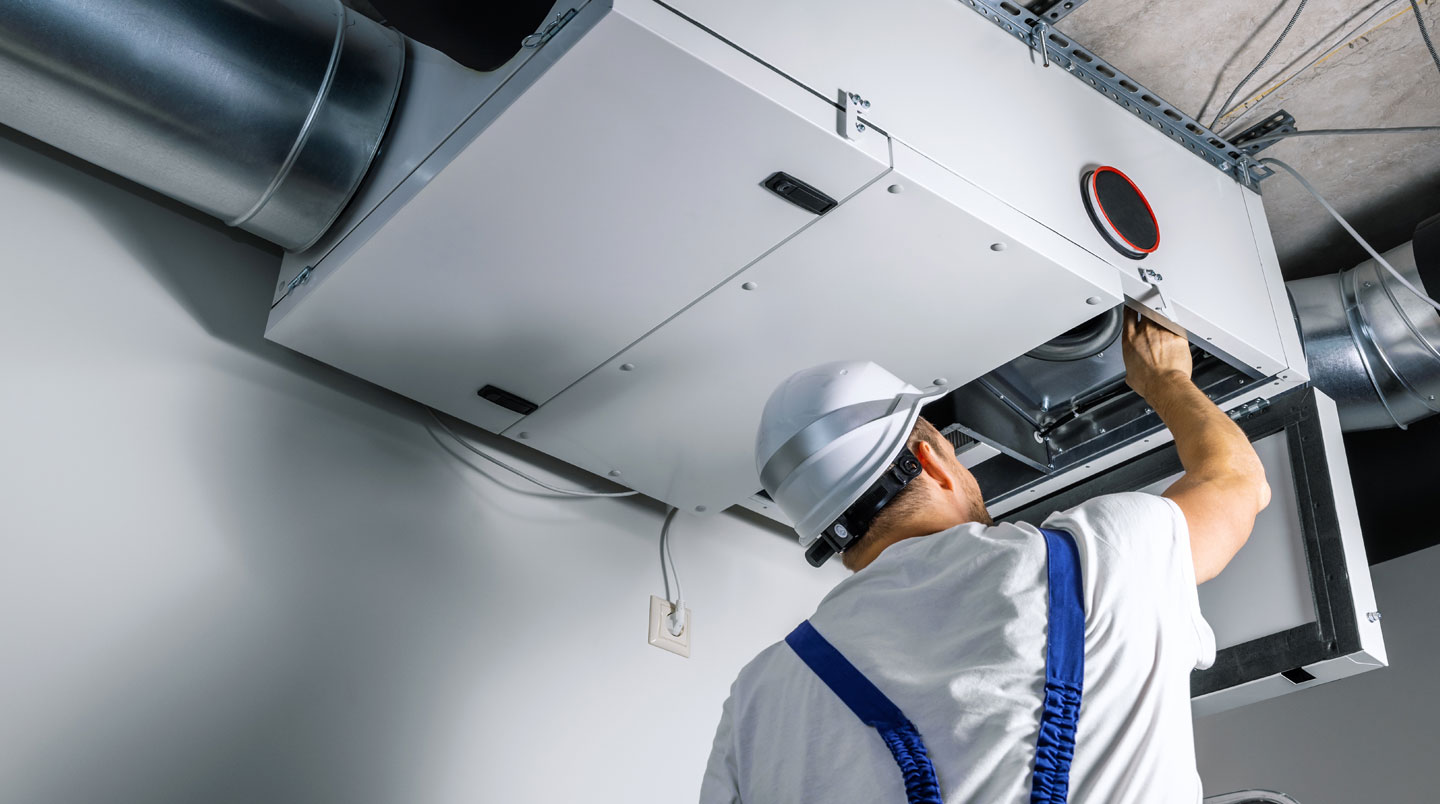Un technicien de maintenance inspecte un système de ventilation industriel en hauteur, vu de dos avec casque de sécurité